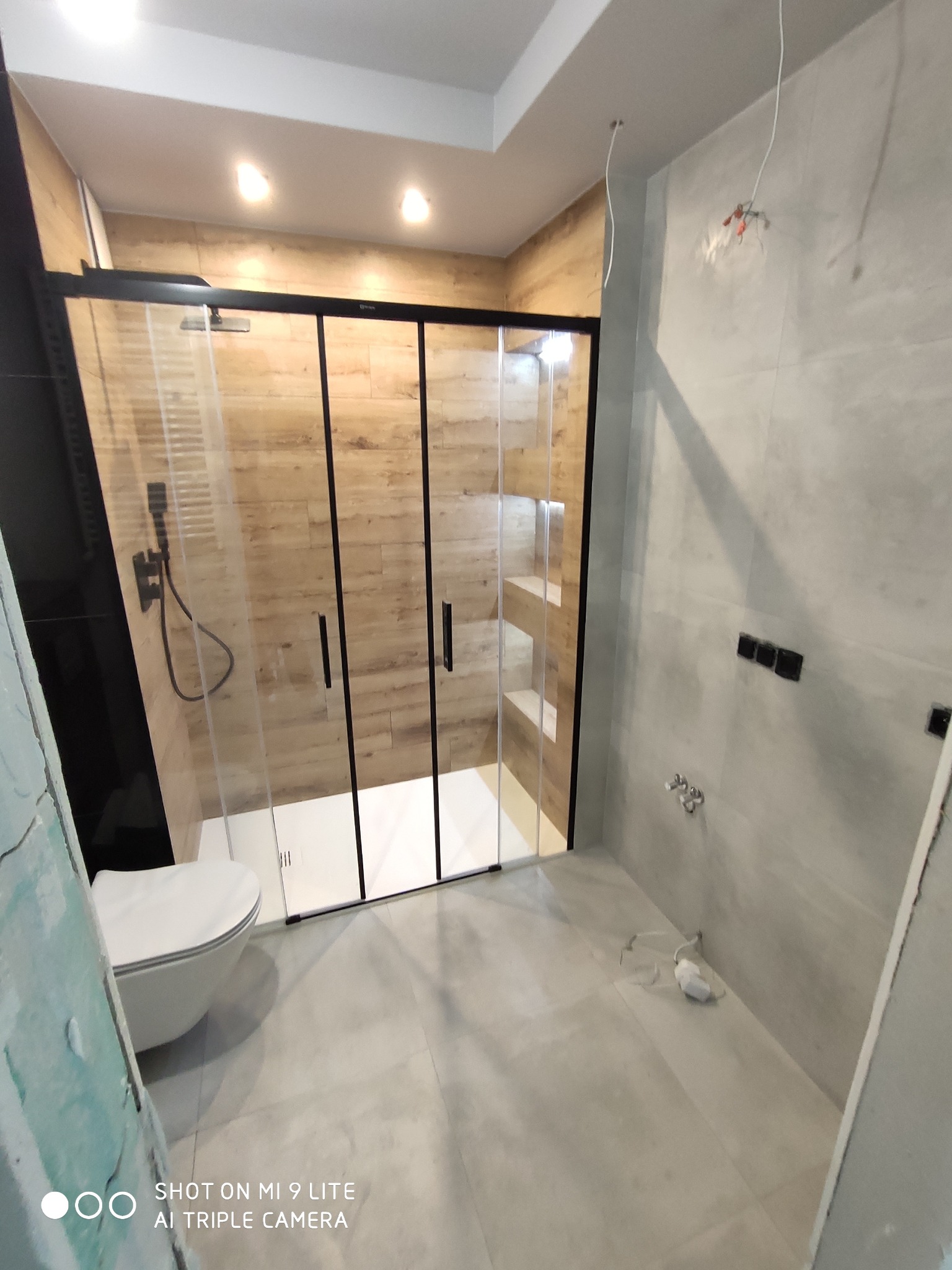 Nowoczesna łazienka w trakcie remontu: kabina prysznicowa z czarnymi ramami i drewnopodobnymi płytkami wewnątrz, toaleta podwieszana, szare płytki na ścianach i podłodze, wystające kable elektryczne.