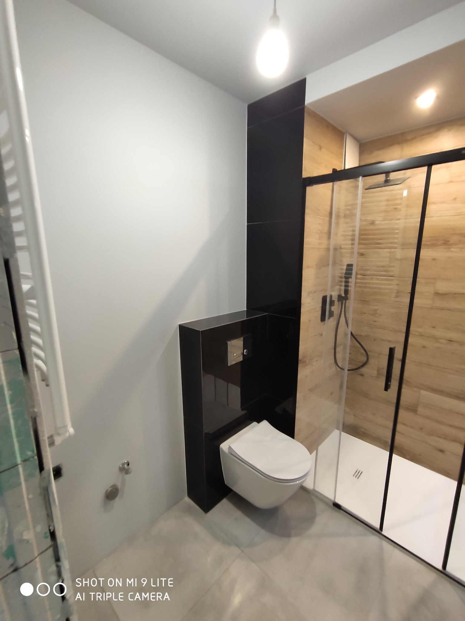 Nowoczesna łazienka z wiszącą toaletą, kabiną prysznicową z czarnymi ramami i drewnopodobnymi płytkami na ścianie, oświetlona ciepłym światłem.