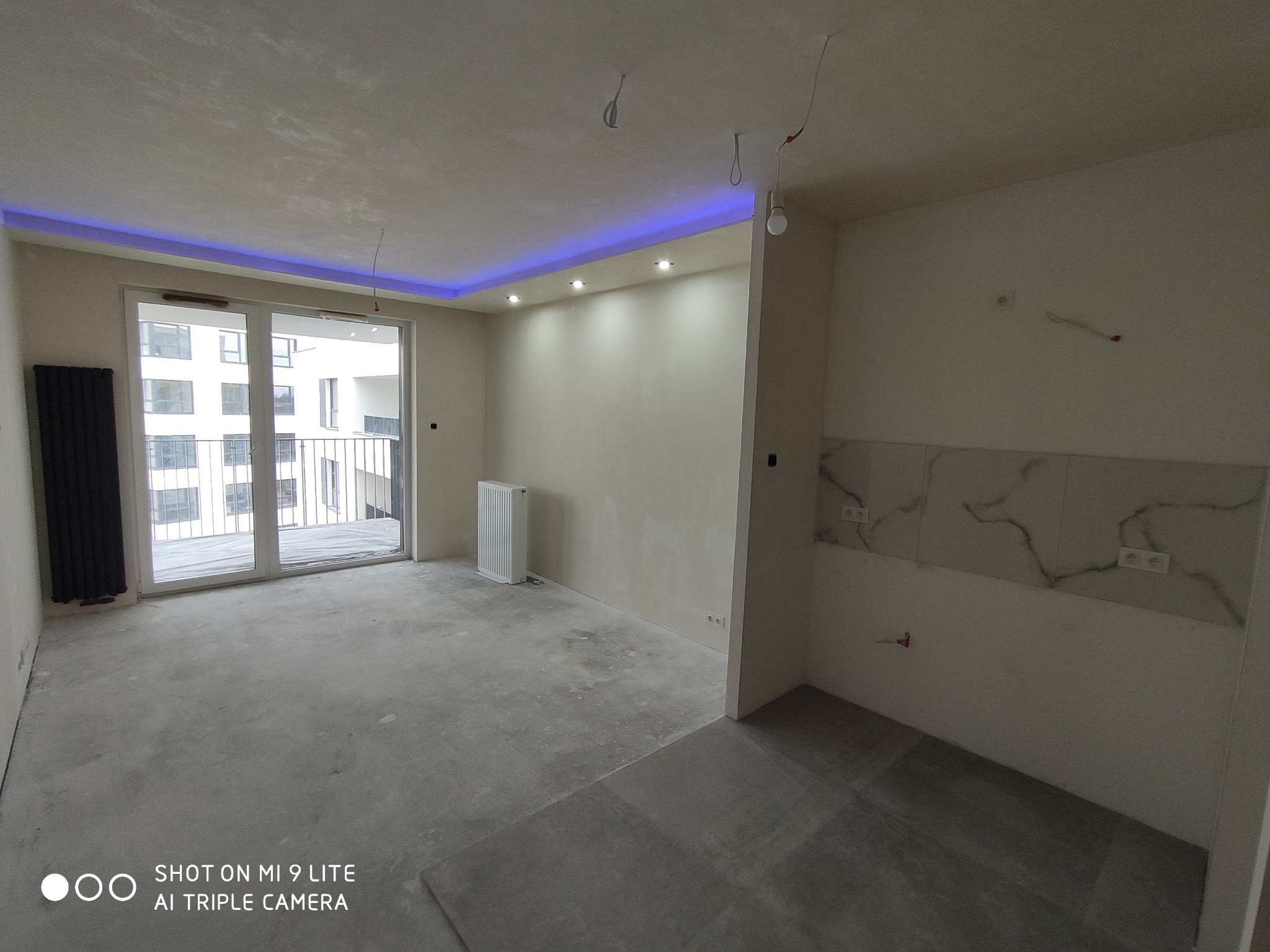 Surowe wnętrze mieszkania w stanie deweloperskim z wylewką, grzejnikiem żeberkowym, grzejnikiem płytowym, marmurową mozaiką na ścianie i podwieszanym sufitem z niebieskim oświetleniem LED.