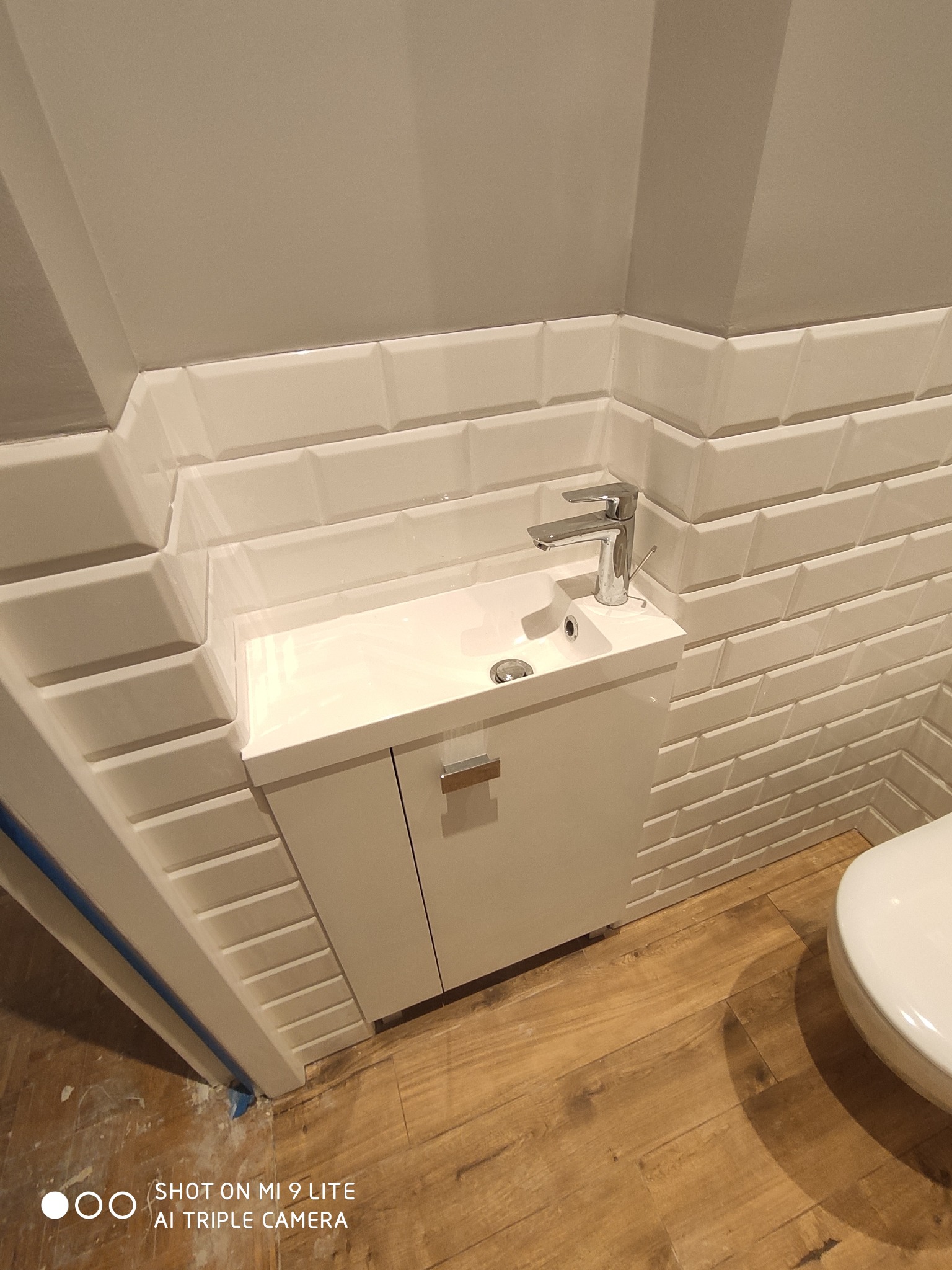 Biała umywalka z szafką w małej łazience, ściany wyłożone białymi, fazowanymi płytkami typu metro, drewnopodobna podłoga.