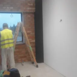 Pracownik w kamizelce odblaskowej stoi przy drabinie w pomieszczeniu z ceglaną ścianą i pomalowaną na biało ścianą, torby robocze na podłodze.