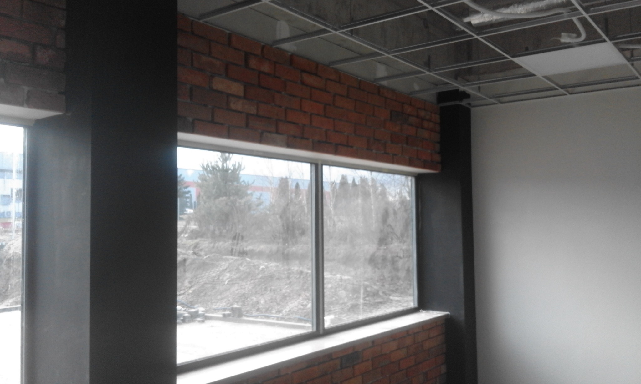 Wnętrze w trakcie remontu: okno w murze z cegły, czarne słupy, sufit z widoczną konstrukcją i częściowo zamontowanymi panelami.