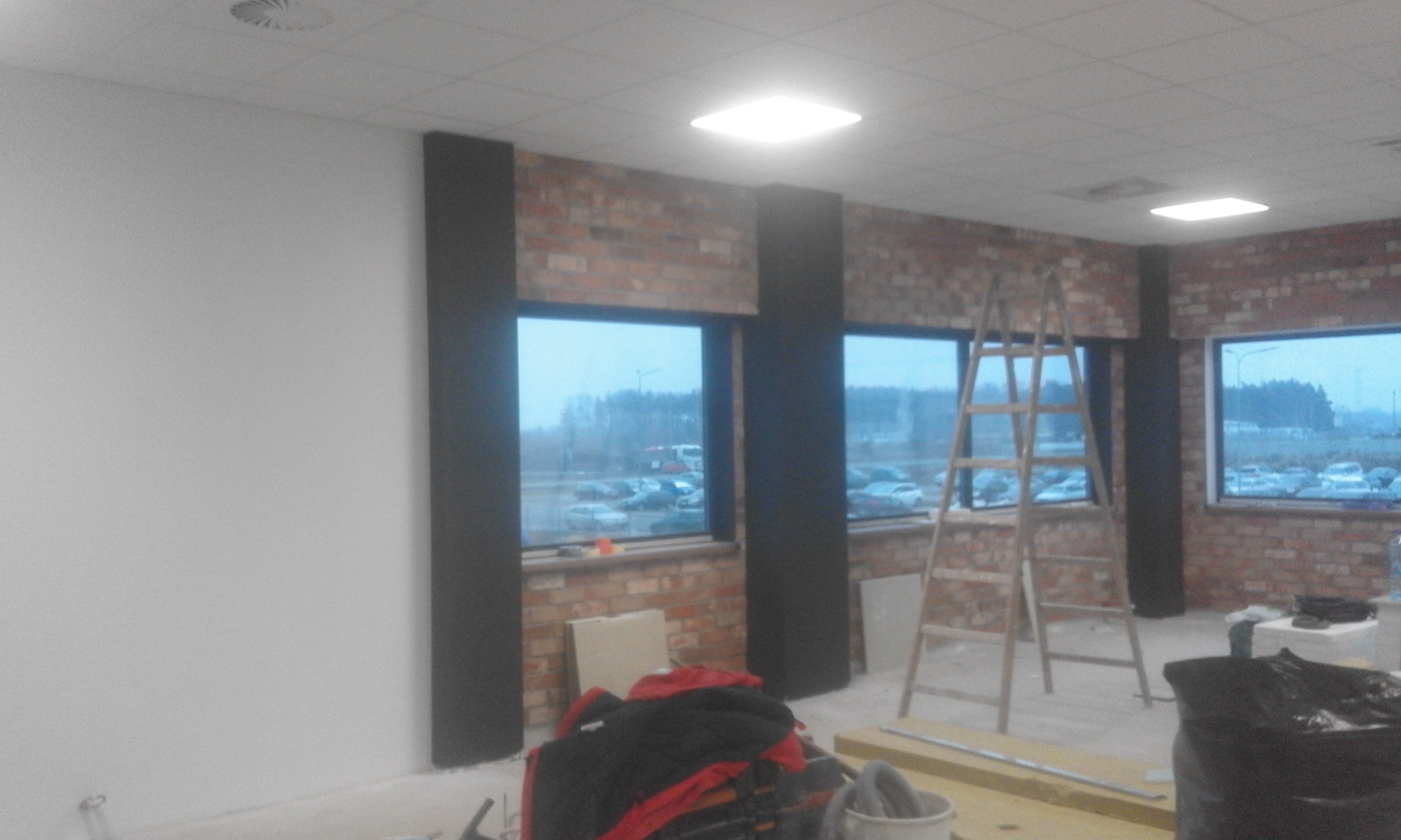 Wnętrze w trakcie prac wykończeniowych: surowa cegła, wstawione okna, drabina oparta o ścianę, narzędzia i materiały budowlane w nieładzie.