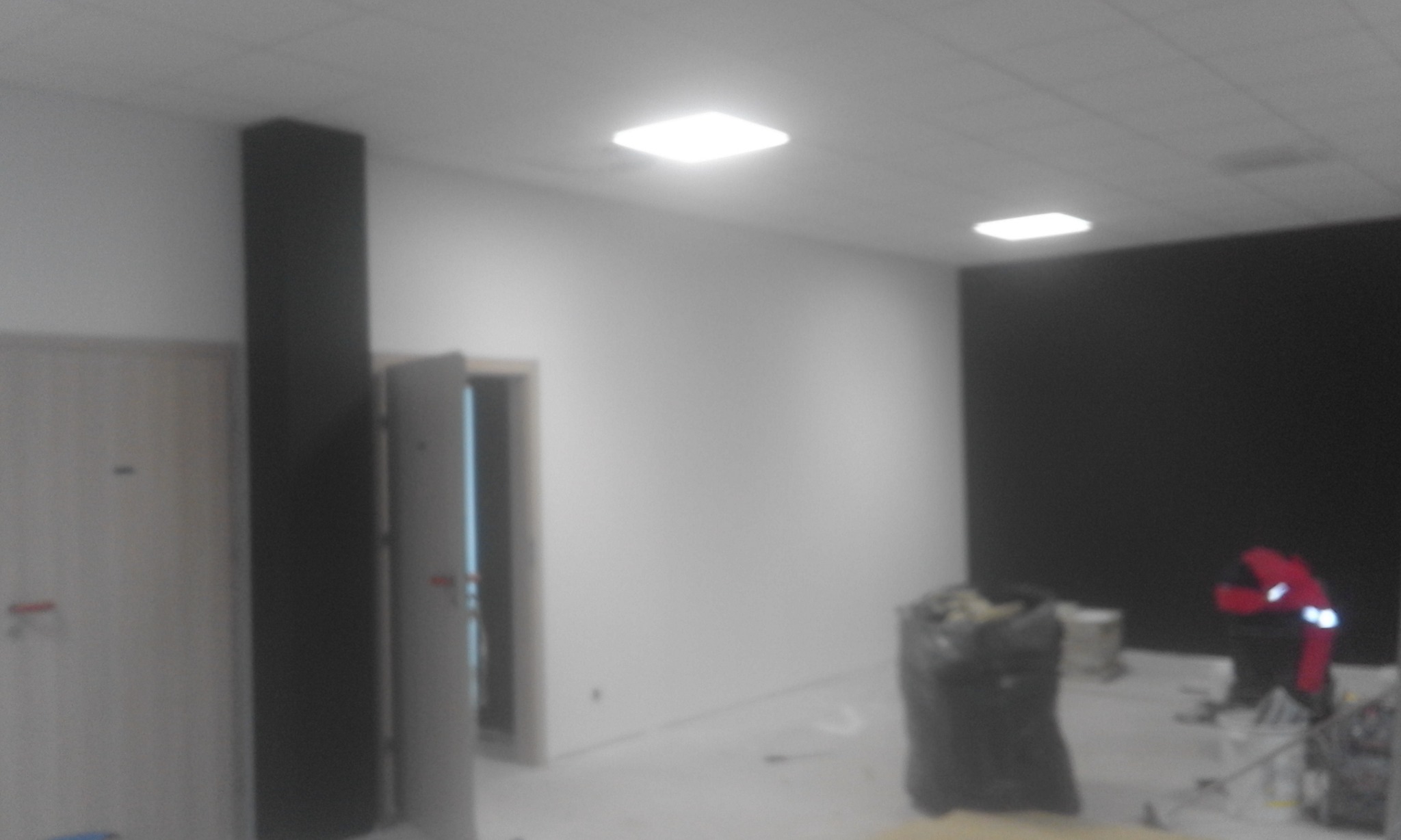 Wnętrze biura w trakcie prac wykończeniowych: ściany częściowo pomalowane na biało i czarno, widoczny pracownik w czerwonym kombinezonie, worki z odpadami i narzędzia.