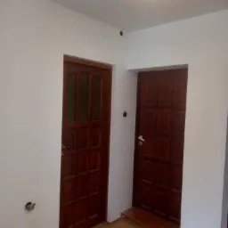 Dwa ciemne, drewniane drzwi wewnętrzne z ościeżnicami w białym pomieszczeniu, jeden z szybkami, widoczny wystający kabel elektryczny w ścianie.