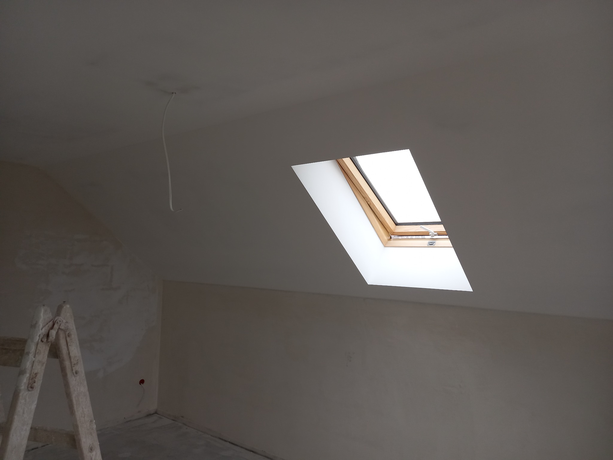 Poddasze w trakcie remontu z oknem dachowym, widoczne ślady po szpachlowaniu i drabina malarska.