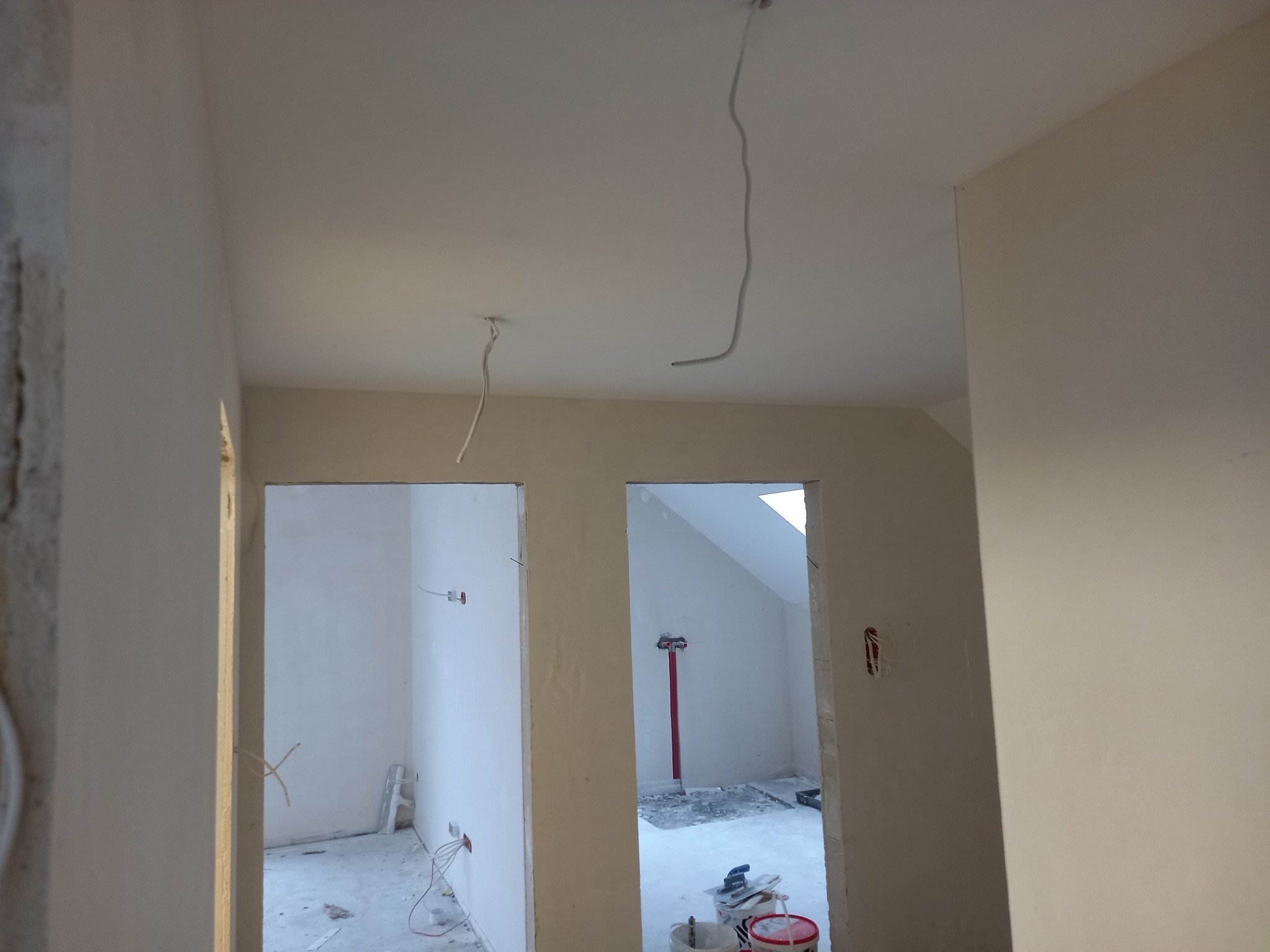 Wnętrze w trakcie prac wykończeniowych: świeżo otynkowane ściany, wystające kable elektryczne, widoczne narzędzia i materiały budowlane w pomieszczeniu obok.