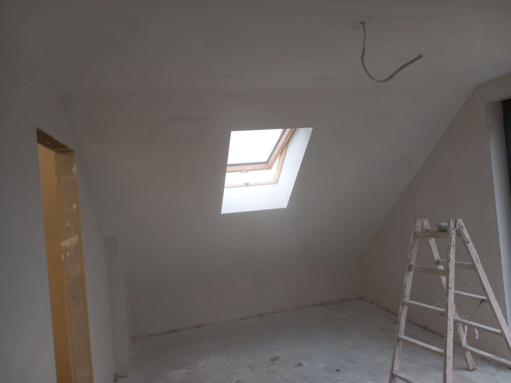 Poddasze w trakcie prac wykończeniowych z oknem dachowym, widoczna drabina malarska, surowy stan ścian przygotowanych do malowania, wystający kabel z sufitu.