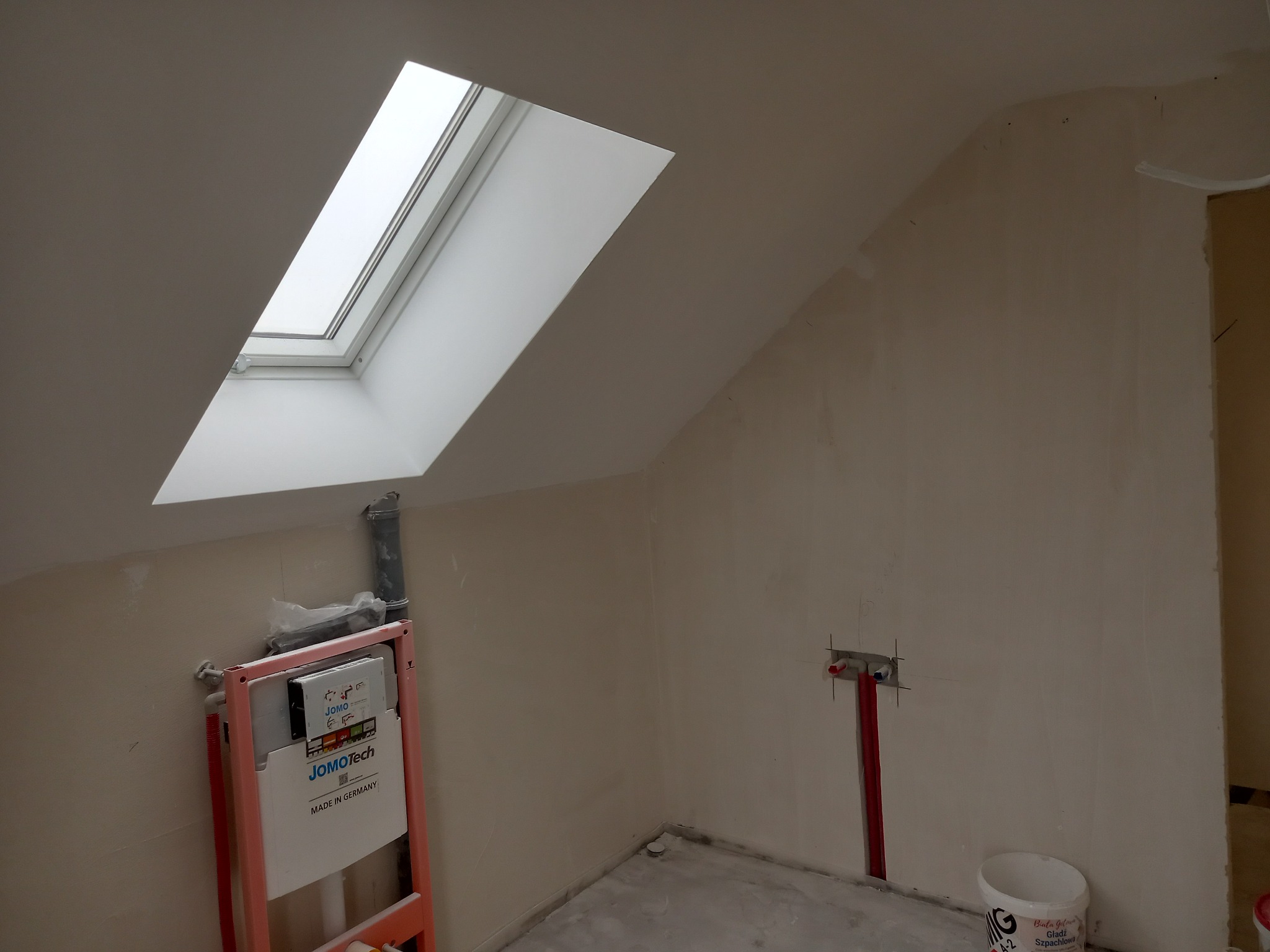 Poddasze w trakcie remontu, widoczne okno dachowe, stelaż podtynkowy WC marki JomoTech, przygotowane instalacje wodne i kanalizacyjne, ściany po szpachlowaniu, gotowe do dalszych prac wykończeniowych.