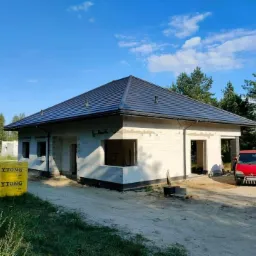 Nowy, ciemny dach na surowym budynku z bloczków Ytong, czerwony samochód dostawczy w tle, błękitne niebo z chmurami.