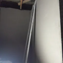 Wnętrze w trakcie prac wykończeniowych, ściana pokryta świeżym tynkiem, widoczne drewniane elementy konstrukcyjne stropu i listwy przypodłogowe opierające się o ścianę.