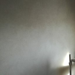 Ap-bud - Świeżo położona, jasnoszara ściana z delikatną fakturą, oświetlona z dołu, z fragmentem rusztowania po prawej stronie. Widoczne surowe wykończenie sufitu.