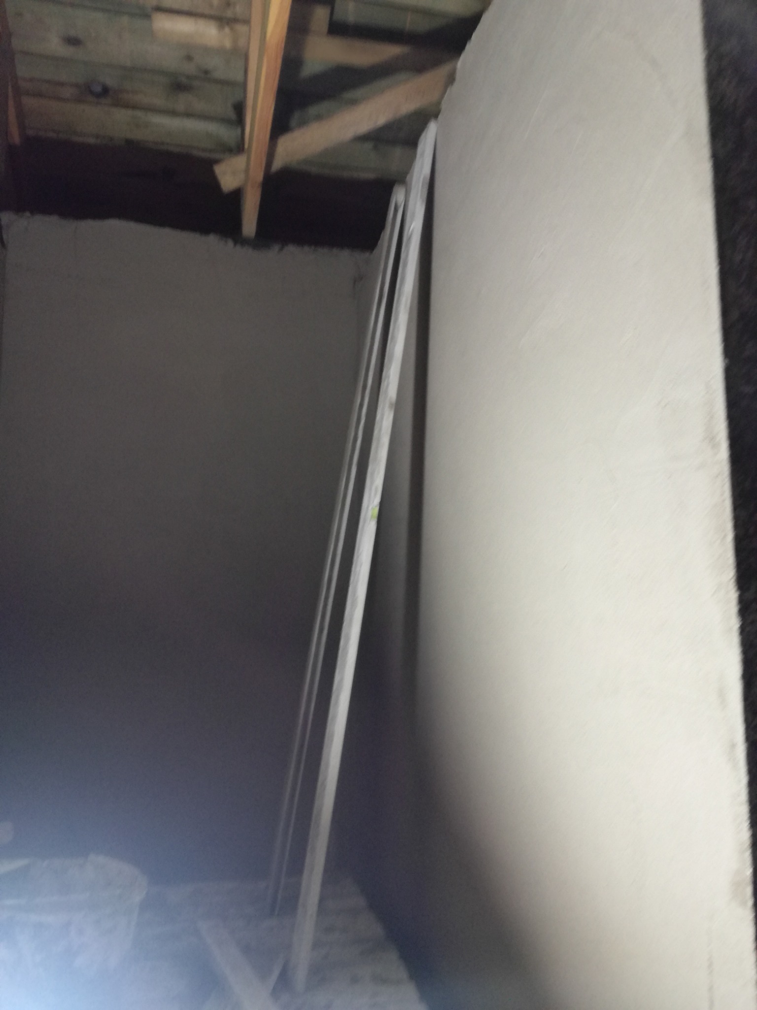 Wnętrze w trakcie prac wykończeniowych, ściana pokryta świeżym tynkiem, widoczne drewniane elementy konstrukcyjne stropu i listwy przypodłogowe opierające się o ścianę.