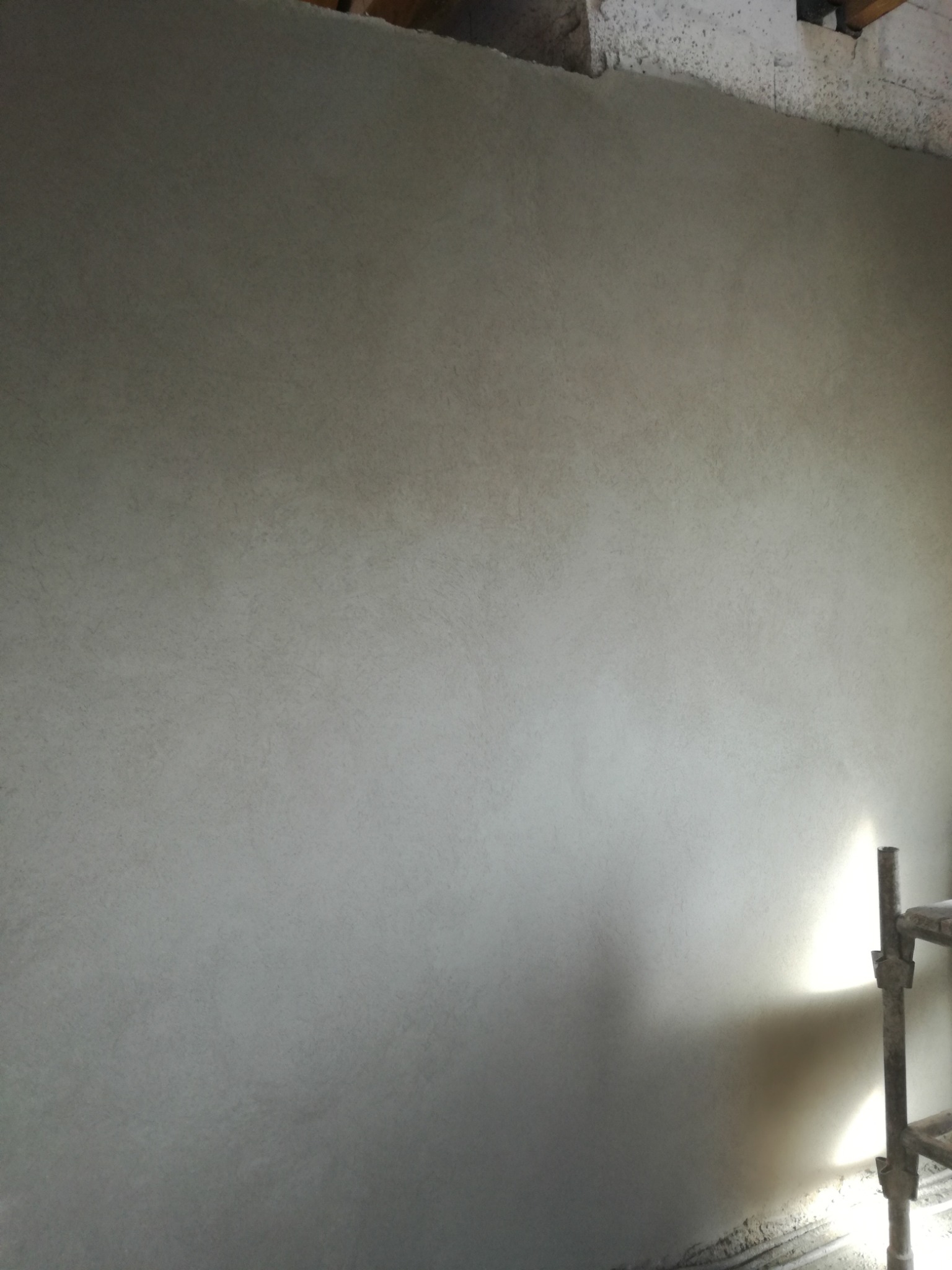 Świeżo położona, jasnoszara ściana z delikatną fakturą, oświetlona z dołu, z fragmentem rusztowania po prawej stronie. Widoczne surowe wykończenie sufitu.