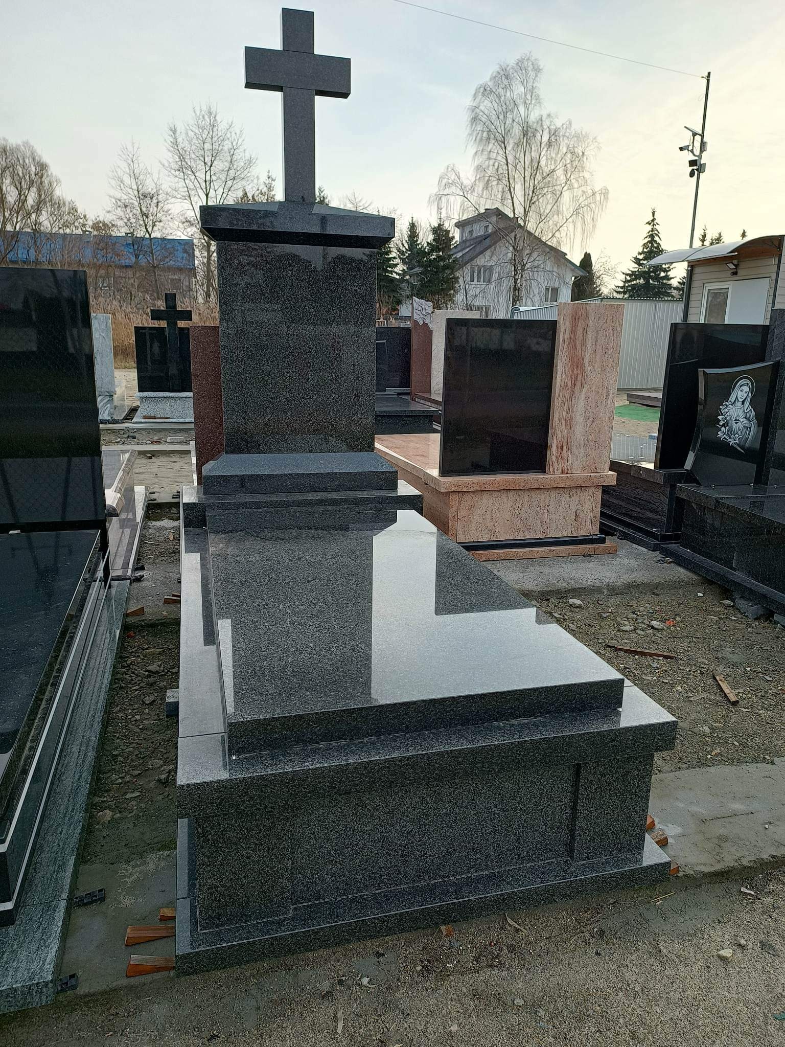 Nowoczesny, ciemny nagrobek z krzyżem na tle innych nagrobków i zabudowań.