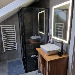Nowoczesna łazienka na poddaszu z kabiną prysznicową typu walk-in, umywalką nablatową na szafce z drewnianymi lamelami, podświetlanym lustrem i grzejnikiem drabinkowym.