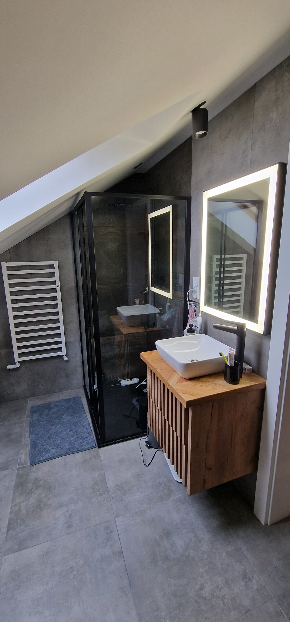 Nowoczesna łazienka na poddaszu z kabiną prysznicową typu walk-in, umywalką nablatową na szafce z drewnianymi lamelami, podświetlanym lustrem i grzejnikiem drabinkowym.