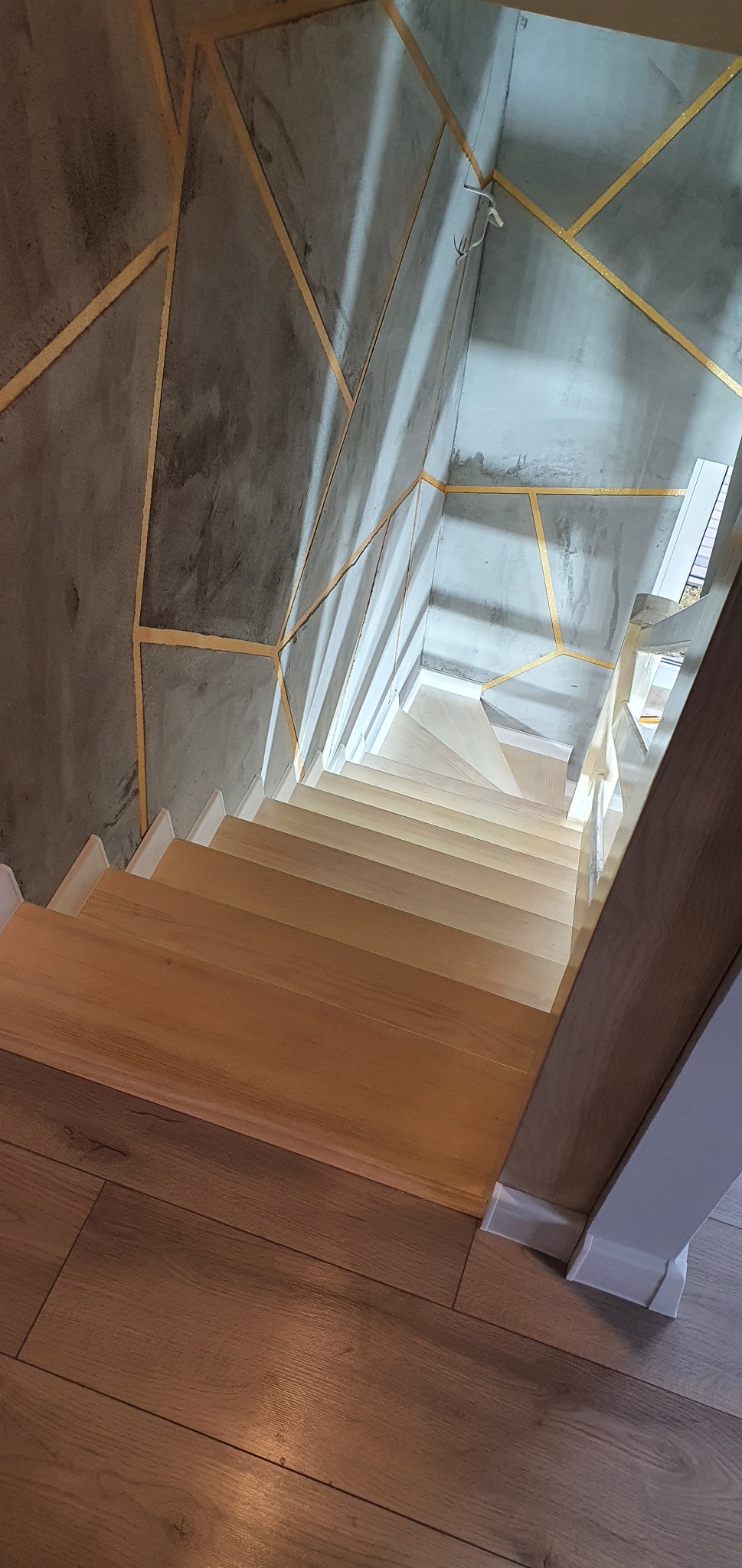 Schody drewniane z betonową ścianą ozdobioną złotymi geometrycznymi wzorami, widok z góry na dół.