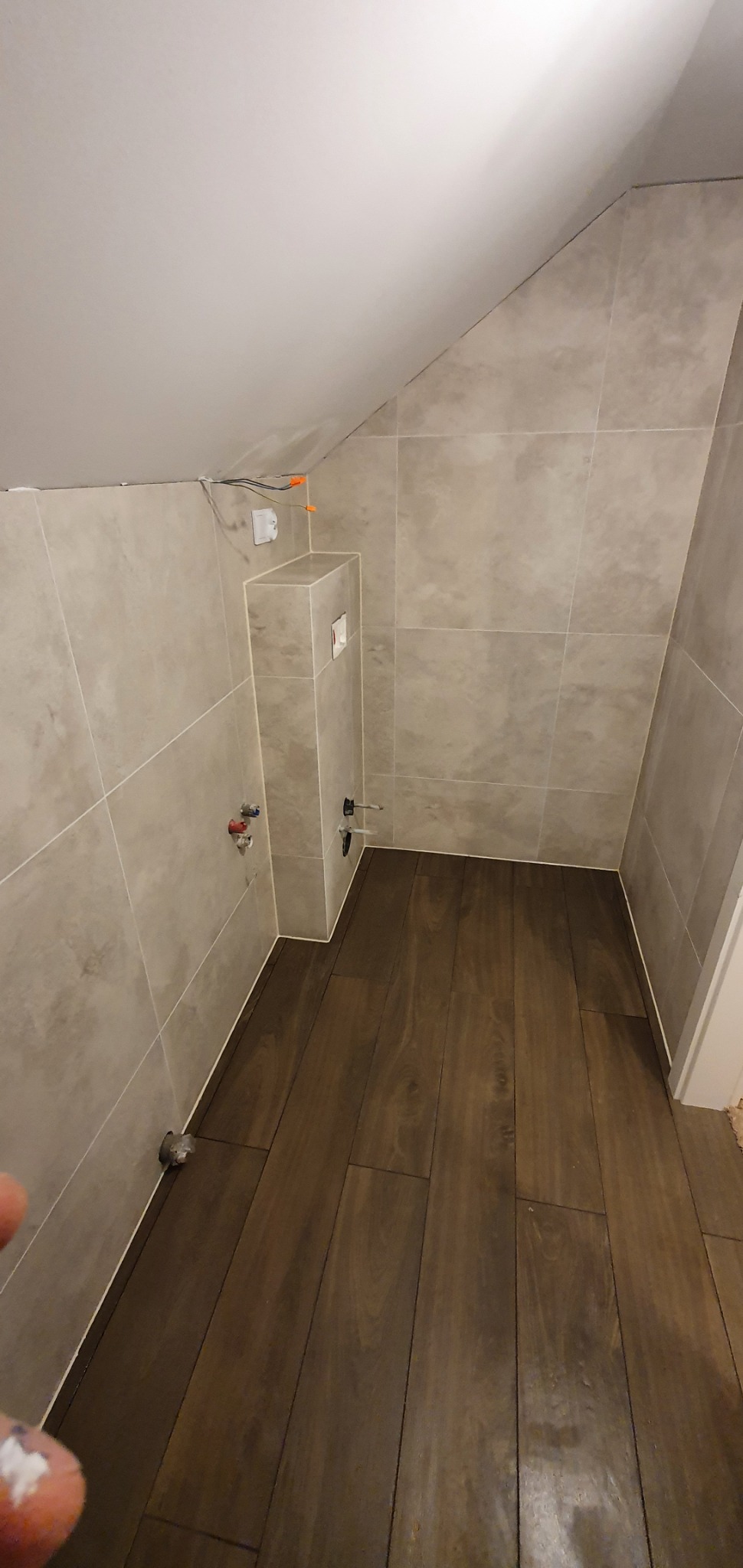 Ukończona łazienka na poddaszu ze skośnym sufitem, wyłożona beżowymi płytkami na ścianach i panelami podłogowymi imitującymi ciemne drewno, widoczne przyłącza wodne i elektryczne.