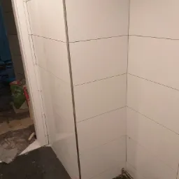 Świeżo ułożone białe, prostokątne płytki z ciemną fugą na ścianie łazienki, widoczne rury instalacji wodnej przy podłodze, w tle resztki materiałów budowlanych i worek z zaprawą.