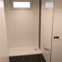 Wnętrze łazienki w Sierakowicach w trakcie wykańczania: białe płytki ścienne, czarna podłoga, zamontowany brodzik i okno, widoczne instalacje wodne i elektryczne przed montażem armatury.