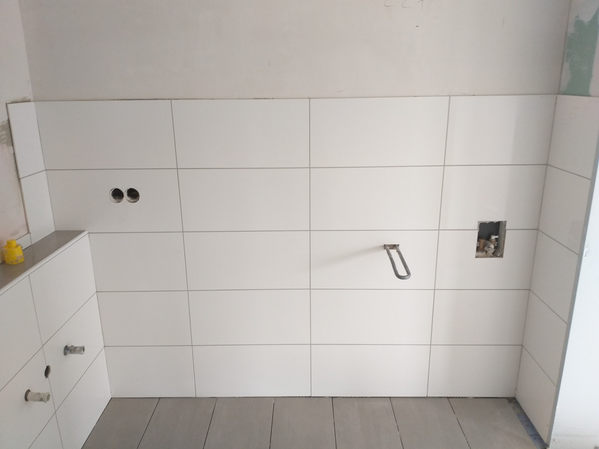 Ściana w trakcie układania białych, prostokątnych płytek w łazience, widoczne otwory na instalacje wodno-kanalizacyjne i gniazdka elektryczne, szare płytki podłogowe, żółty pojemnik z klejem w rogu.