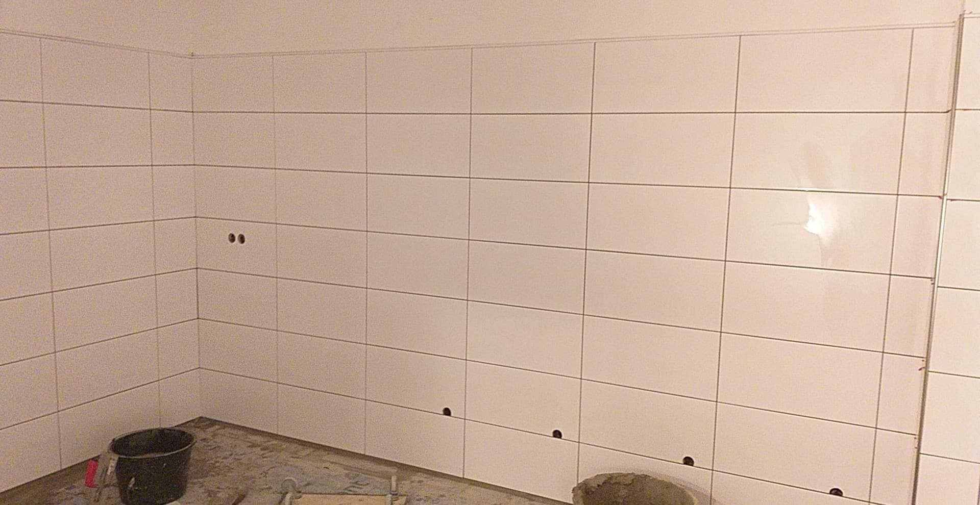 Ściana w trakcie układania białych, prostokątnych płytek ceramicznych w łazience, widoczne otwory na instalacje, wiadro i narzędzia na podłodze.