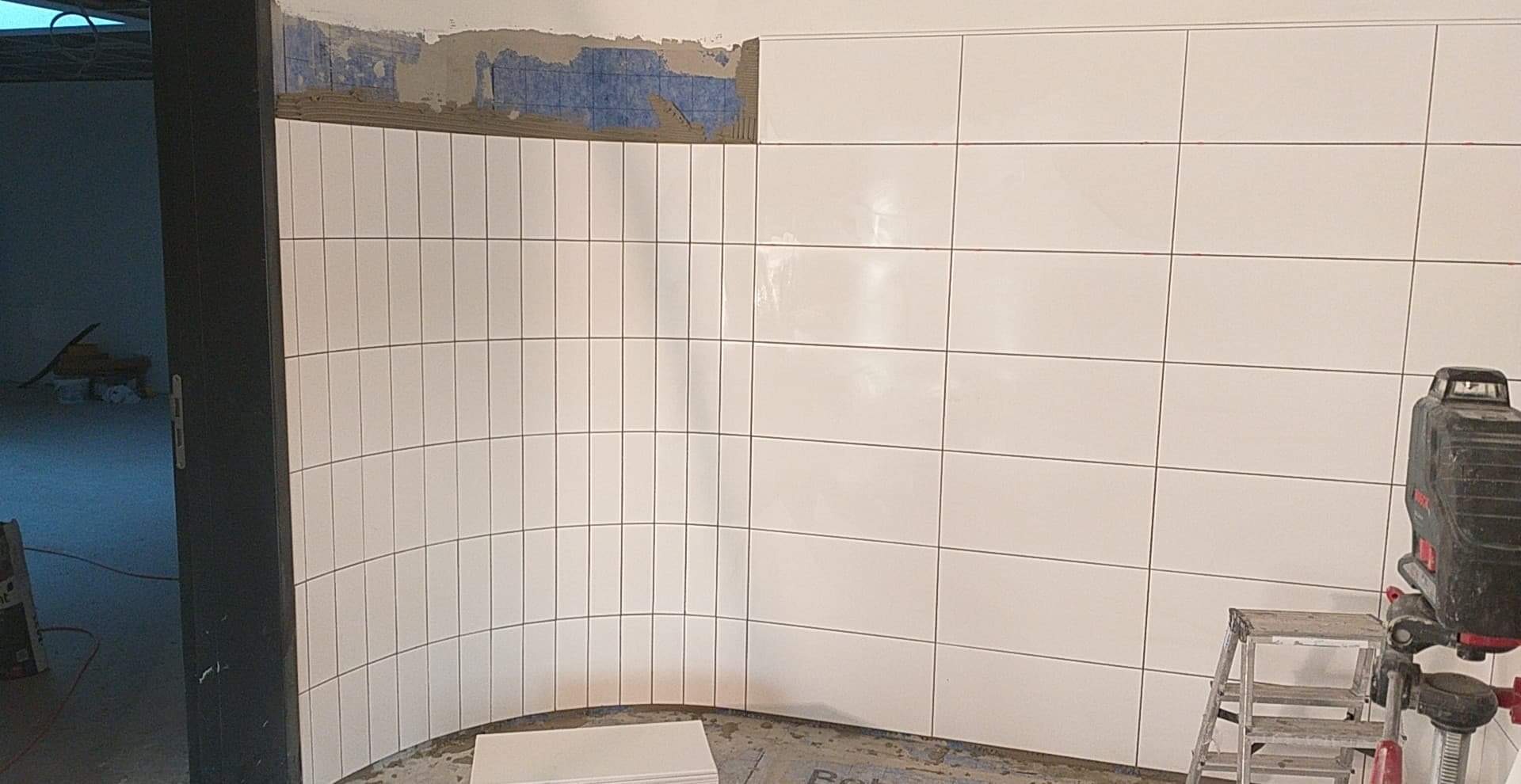 Białe, prostokątne płytki ułożone na zakrzywionej ścianie, widoczny laser krzyżowy na statywie i fragment niezakończonej ściany z resztkami kleju.