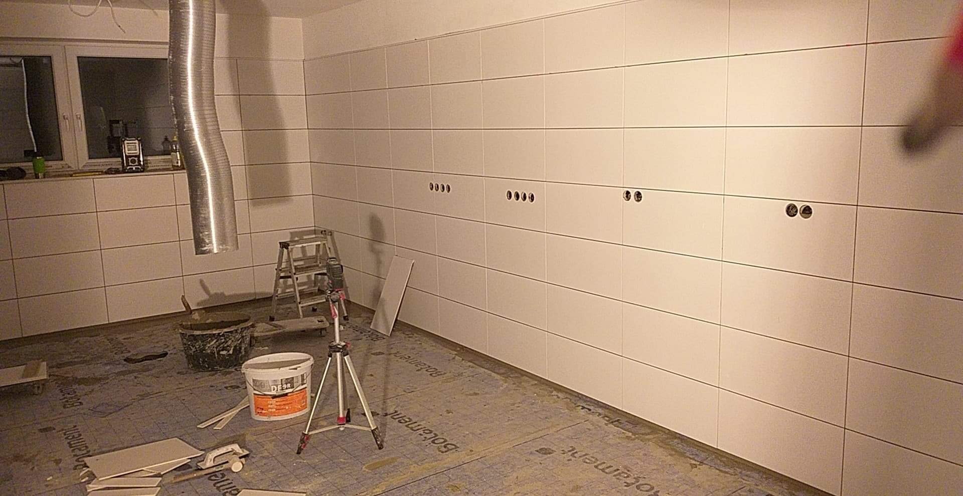 Świeżo wykafelkowana ściana w trakcie prac wykończeniowych; widoczne puszki elektryczne, drabina, wiadro z zaprawą oraz zabezpieczona folią podłoga.