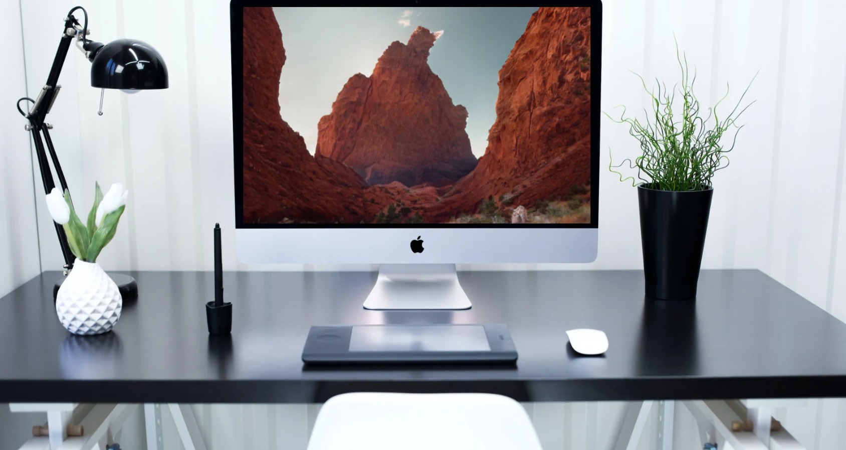 Stanowisko pracy z komputerem iMac wyświetlającym krajobraz górski, tablet graficzny, mysz, lampka biurkowa, wazon z tulipanami i doniczka z rośliną na czarnym biurku.