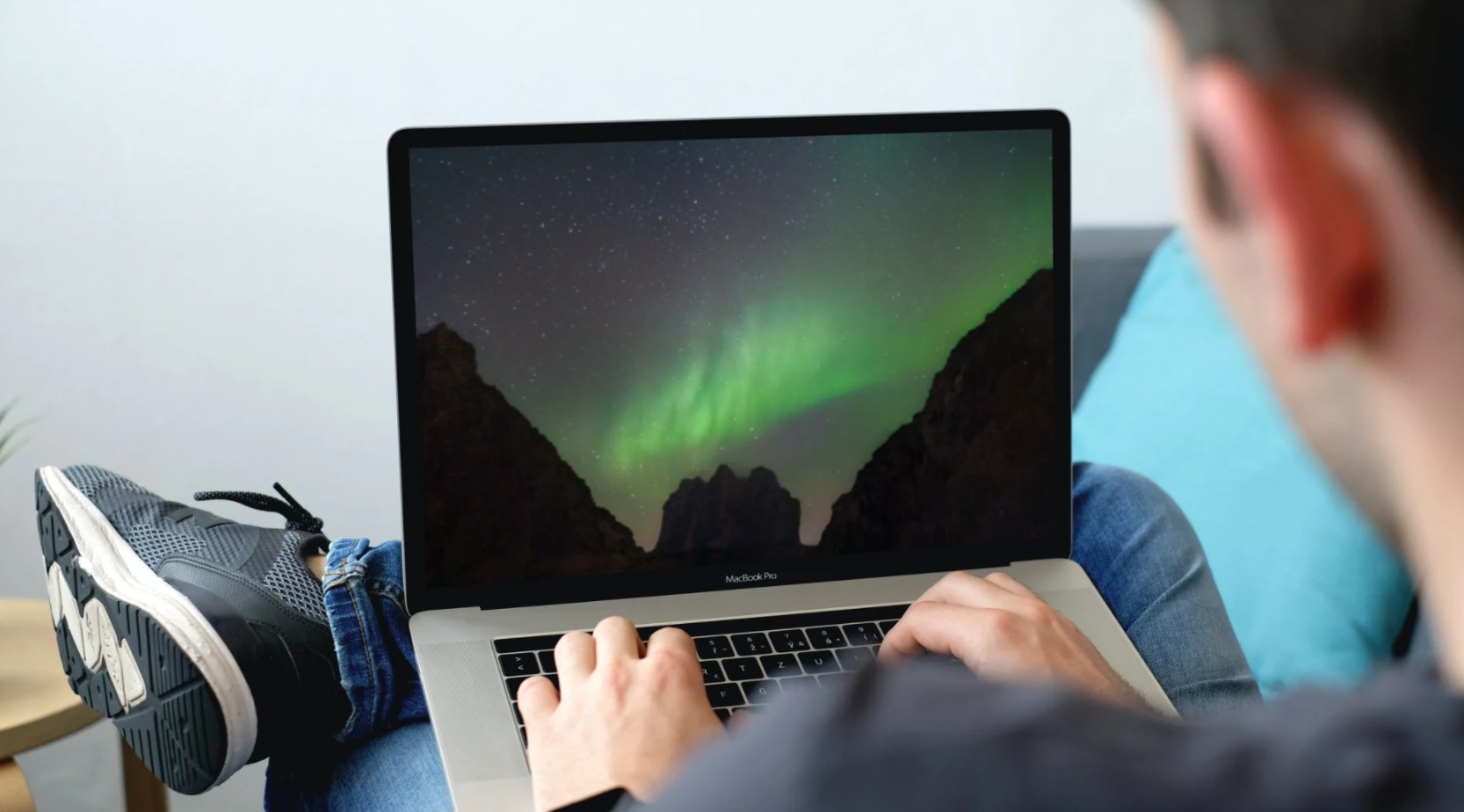 Mężczyzna siedzi z laptopem MacBook Pro, na ekranie wyświetla się zorza polarna między górami, na nogach ma sportowe buty.