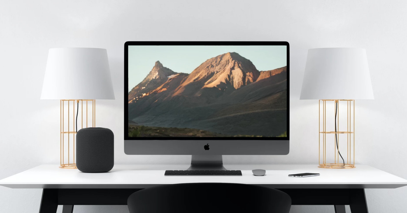 Minimalistyczne biurko z komputerem iMac wyświetlającym górski krajobraz, otoczone lampami z ażurowymi złotymi podstawami i głośnikiem HomePod, całość na tle białej ściany.