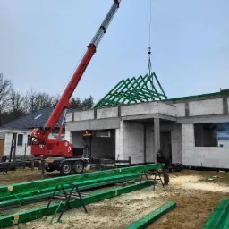 Montaż zielonej więźby dachowej za pomocą czerwonego dźwigu na budowie domu jednorodzinnego; widoczne przygotowane elementy więźby i pracownicy.