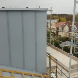 Szary dach z blachy na budynku, widoczny z rusztowania. Metalowe panele, drewniana drabina i fragment instalacji odgromowej. W tle zabudowa mieszkalna.