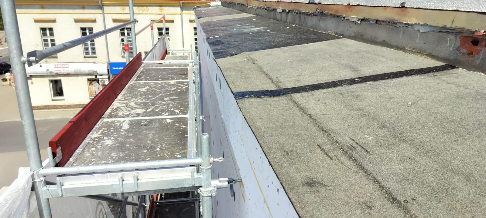 Widok z rusztowania na fragment płaskiego dachu pokrytego papą, z widocznymi łatami i izolacją termiczną ściany attyki.