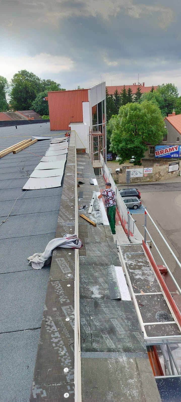Prace dekarskie na dachu budynku z widocznym pracownikiem montującym elementy izolacyjne na krawędzi dachu, z wykorzystaniem rusztowania i poziomicy.