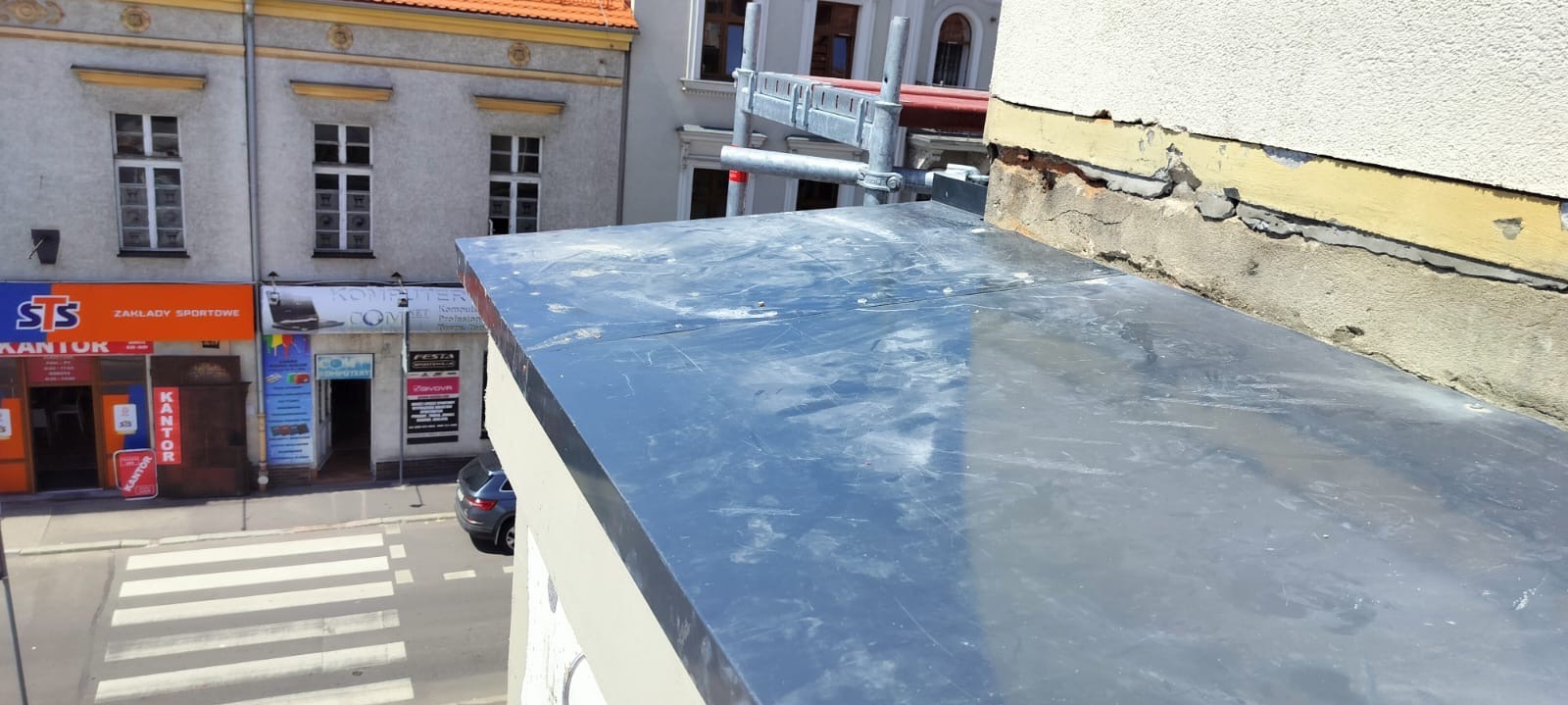 Szare, metalowe wykończenie dachu z widocznymi śladami użytkowania i zarysowaniami, z fragmentem rusztowania i uszkodzoną elewacją budynku w tle, widok z góry na ulicę miejską.