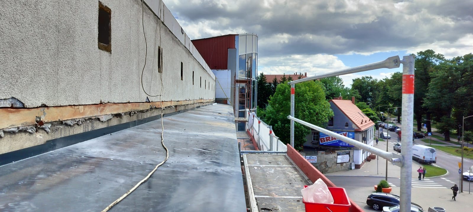 Przygotowania do prac na dachu budynku w Łagiewnikach: widoczna płaska powierzchnia dachu pokryta papą, ściana budynku z ubytkami tynku oraz fragment rusztowania z widokiem na okoliczną zabudowę.