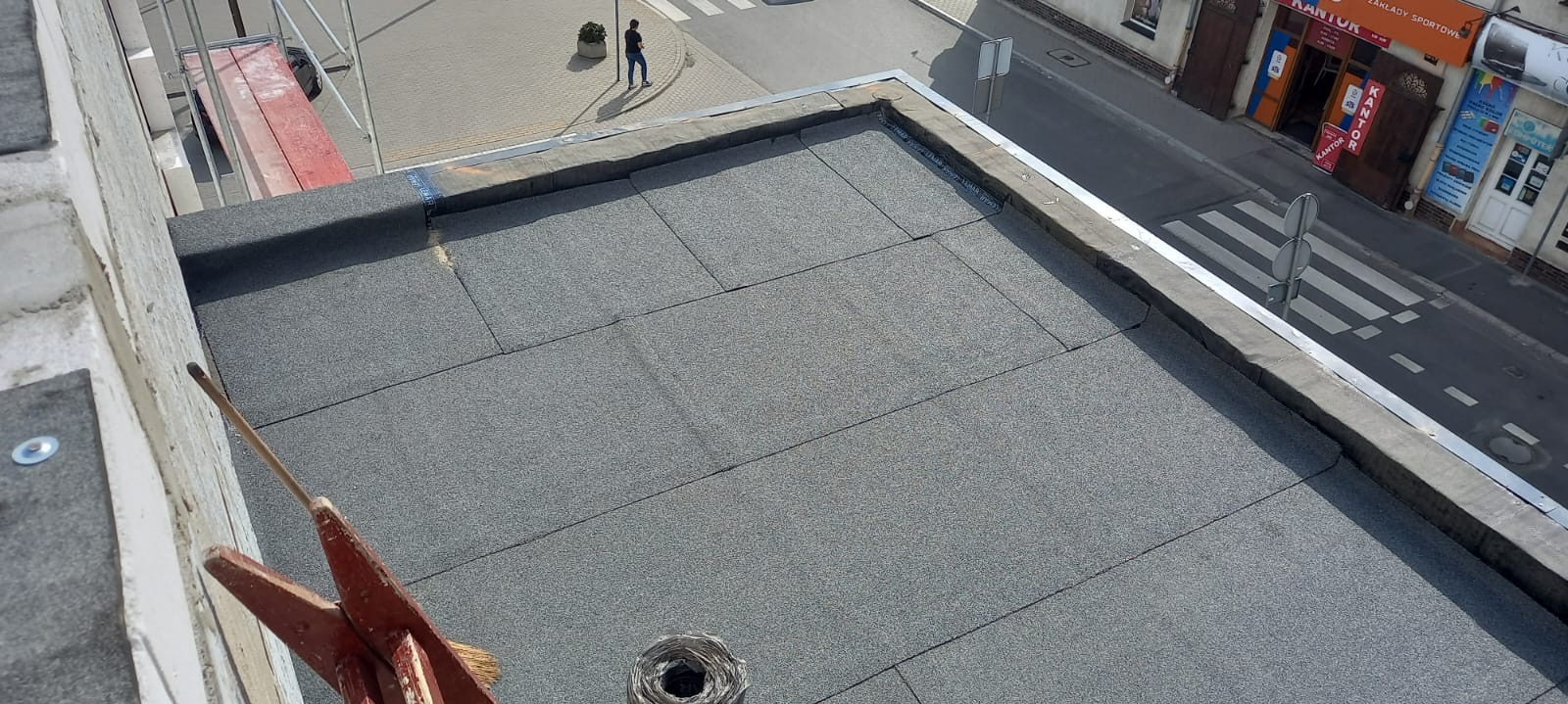 Widok z góry na płaski dach pokryty papą termozgrzewalną, z widocznym odpływem, obróbką blacharską na krawędzi i fragmentem rusztowania przy ścianie budynku. W tle ulica z przejściem dla pieszych...