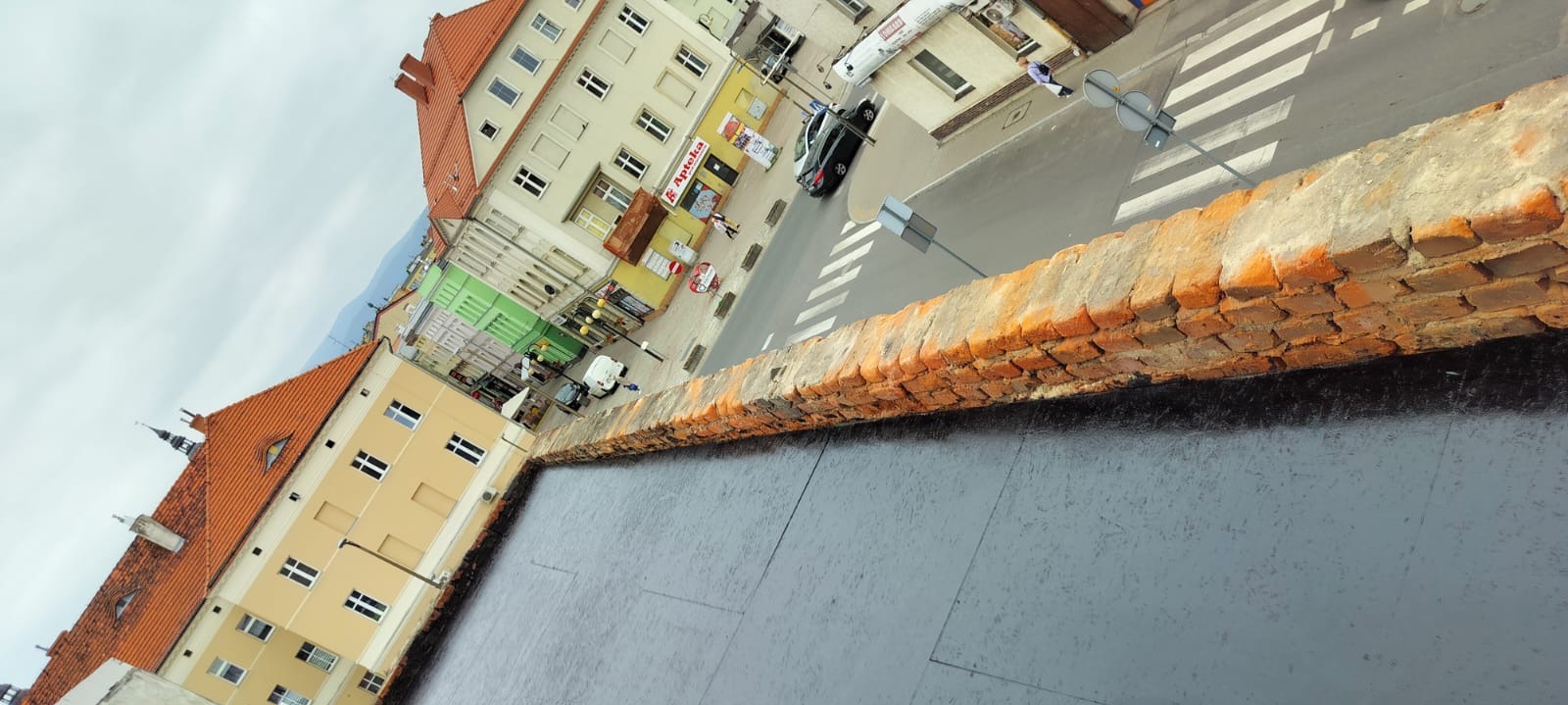 Widok z góry na czarną, płaską powierzchnię dachu z ceglanym murem attykowym w tle, z widokiem na ulicę, budynki i przechodniów w oddali.