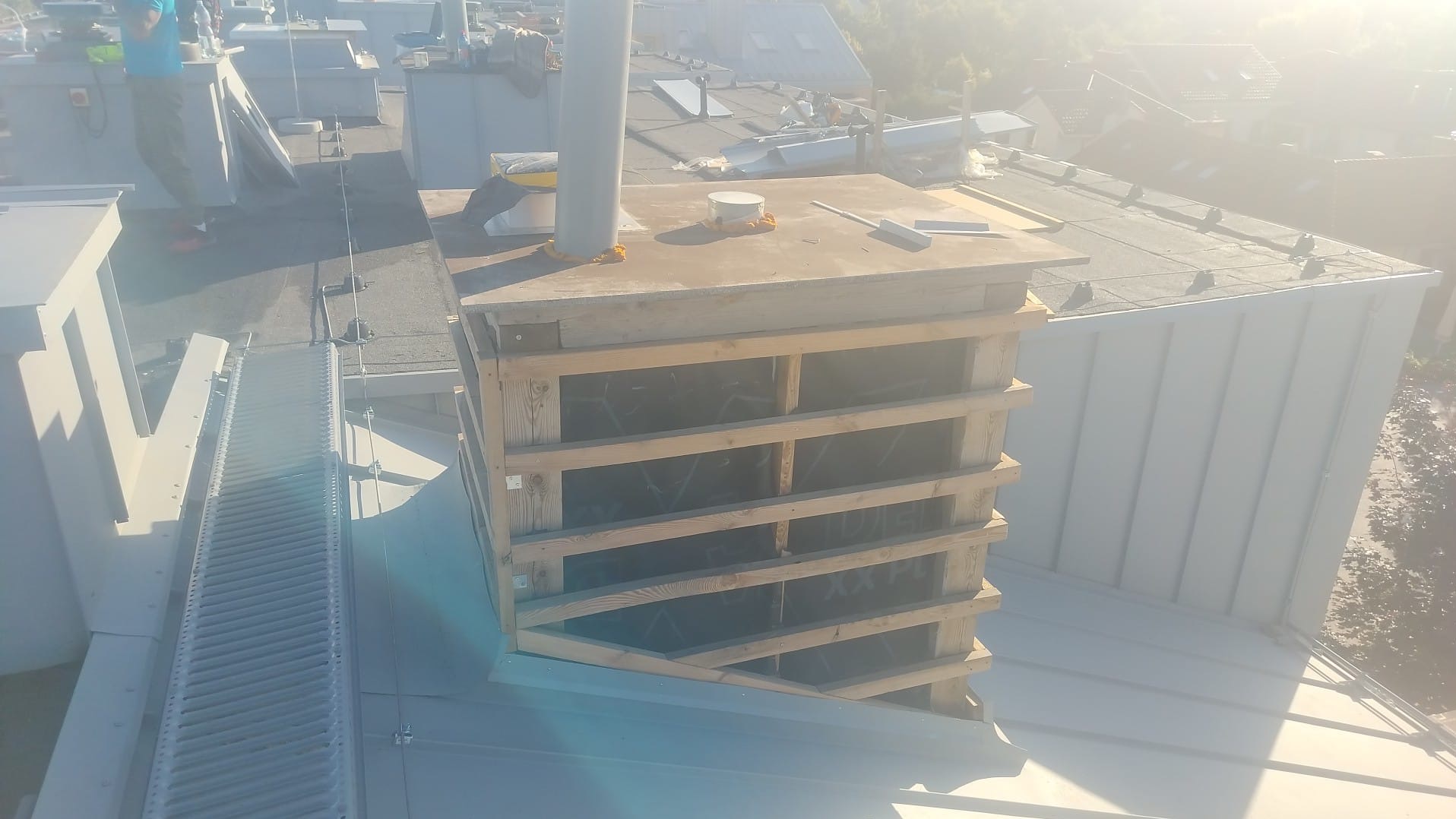 Konstrukcja drewniana na dachu budynku, prawdopodobnie obudowa komina lub innego elementu instalacji dachowej, widok z góry, w tle zabudowa miejska.