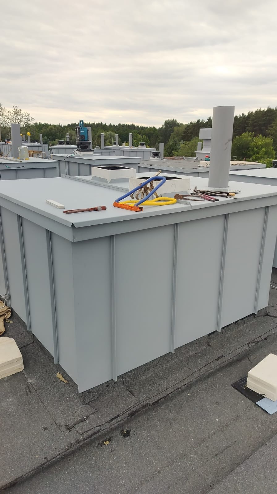 Szare, metalowe obudowy na dachu pokrytym papą, z narzędziami blacharskimi na wierzchu. W tle drzewa i niebo.