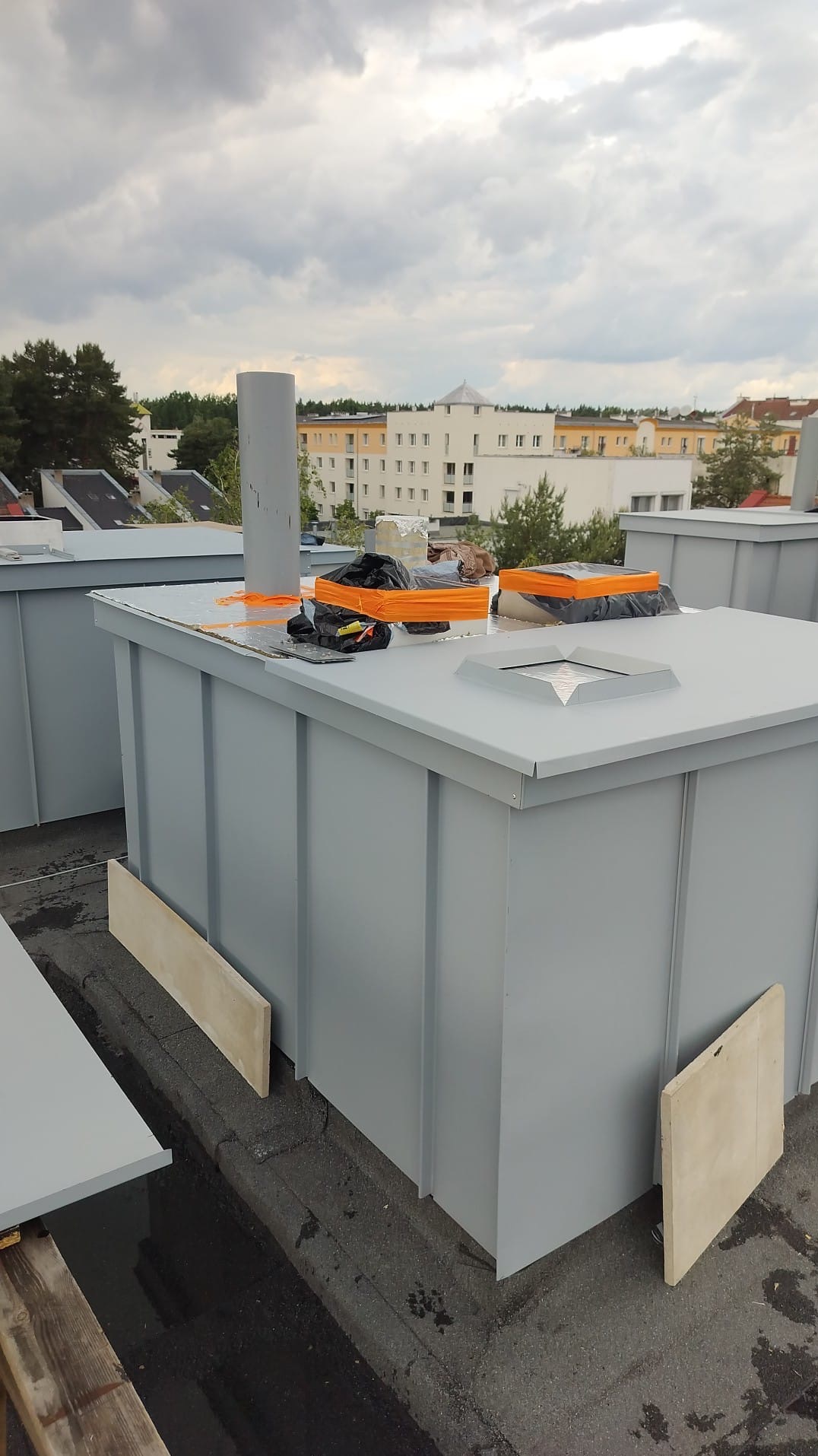 Szare, metalowe obudowy na dachu budynku, prawdopodobnie wentylacyjne, z widocznymi elementami montażowymi i narzędziami. W tle zabudowa miejska i pochmurne niebo.