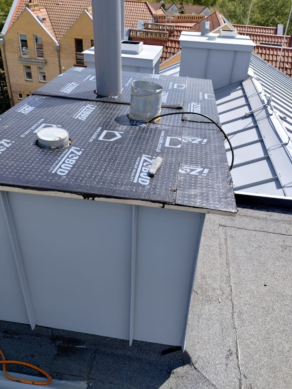 Szczegół dachu z papą izolacyjną Izobud, widoczny komin i metalowe elementy. Ujęcie z góry na płaską powierzchnię dachu, w tle budynki z czerwonymi dachówkami.