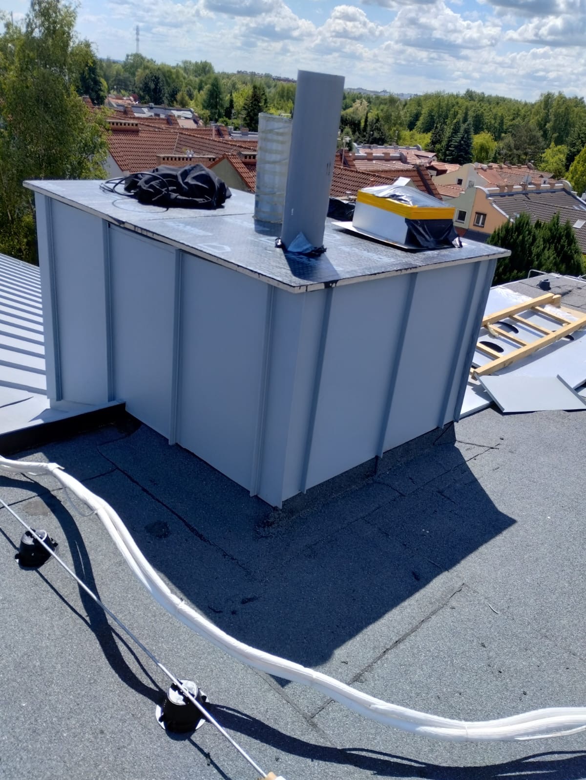 Szare obłożenie komina na dachu z papy, widoczne elementy wentylacyjne i przewody. W tle zabudowa miejska i zieleń, jasne niebo.