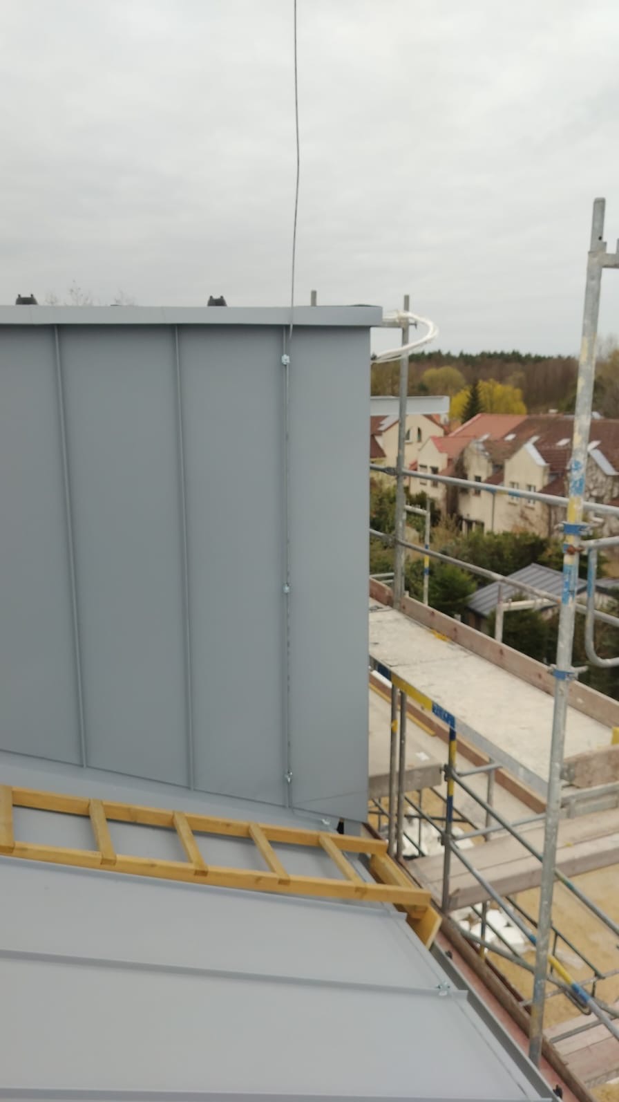 Szary dach z blachy na budynku, widoczny z rusztowania. Metalowe panele, drewniana drabina i fragment instalacji odgromowej. W tle zabudowa mieszkalna.