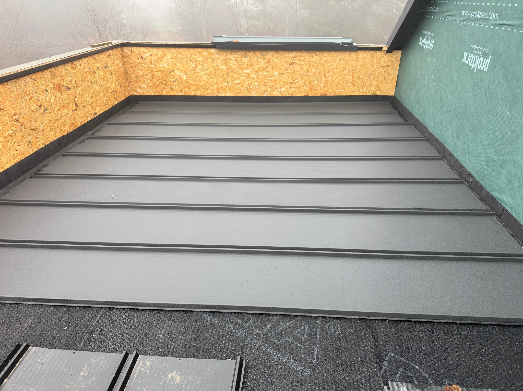 Szare, metalowe panele dachowe ułożone na czarnej membranie, widoczne fragmenty drewnianej konstrukcji i zielonej membrany paroprzepuszczalnej. Detal pokrycia dachu.