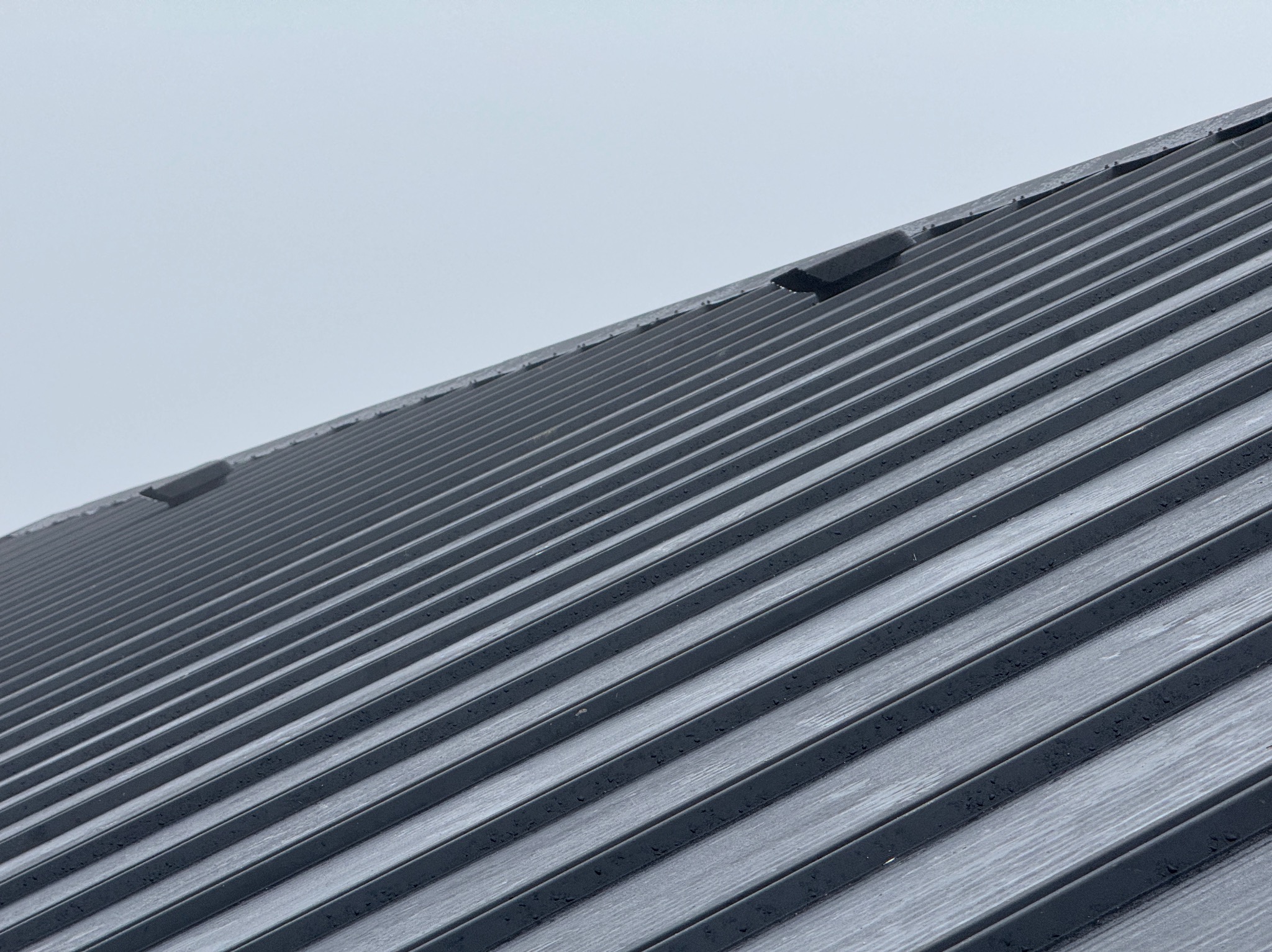 Szary dach z blachy trapezowej pod pochmurnym niebem, widoczny z dołu pod kątem. Charakterystyczne, regularne przetłoczenia blachy. Fragment dachu z obróbką blacharską.