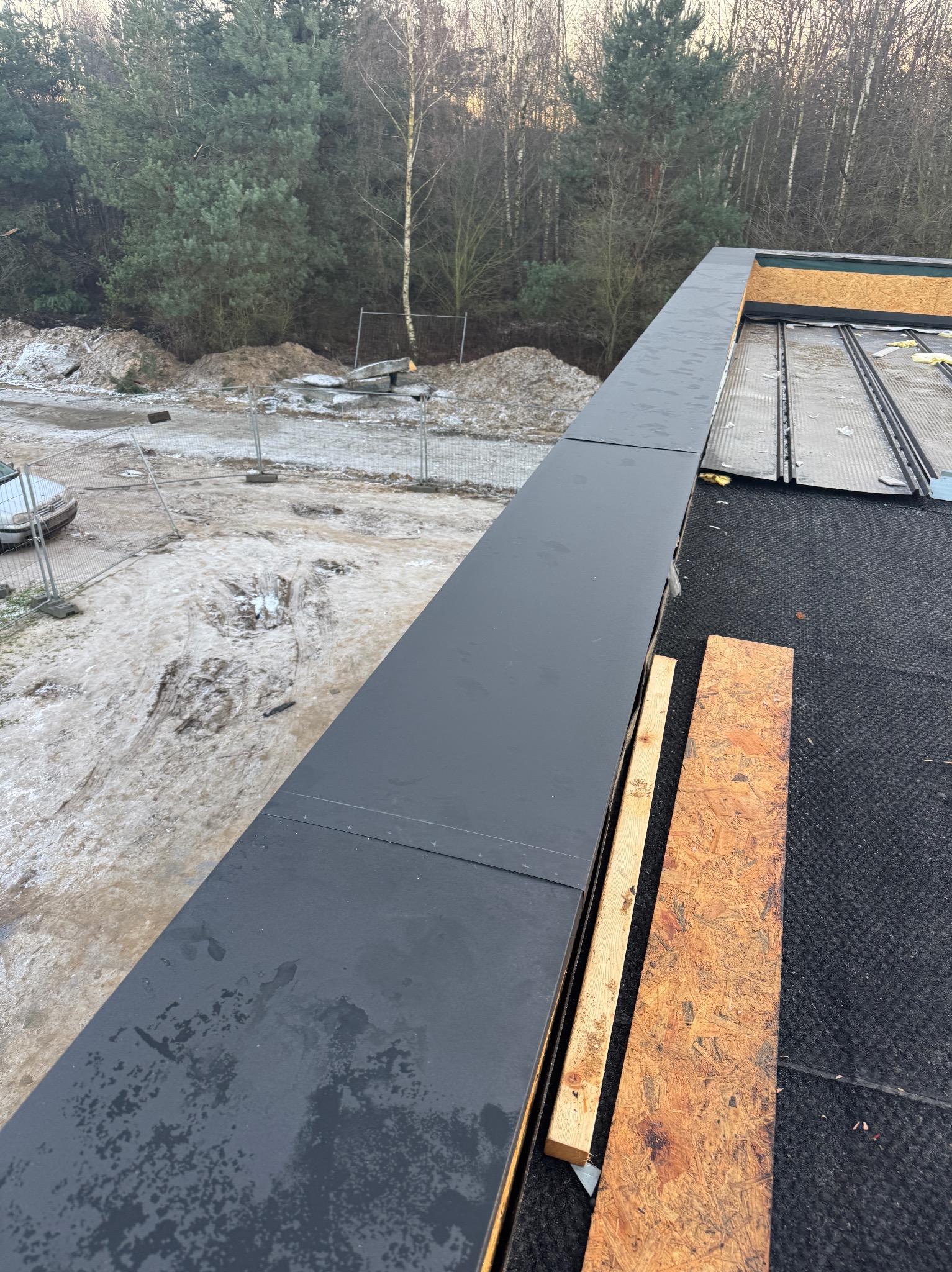 Czarna, metalowa obróbka blacharska na dachu płaskim z widocznymi elementami konstrukcyjnymi i fragmentem zimowego krajobrazu w tle. Detal wykończenia dachu.