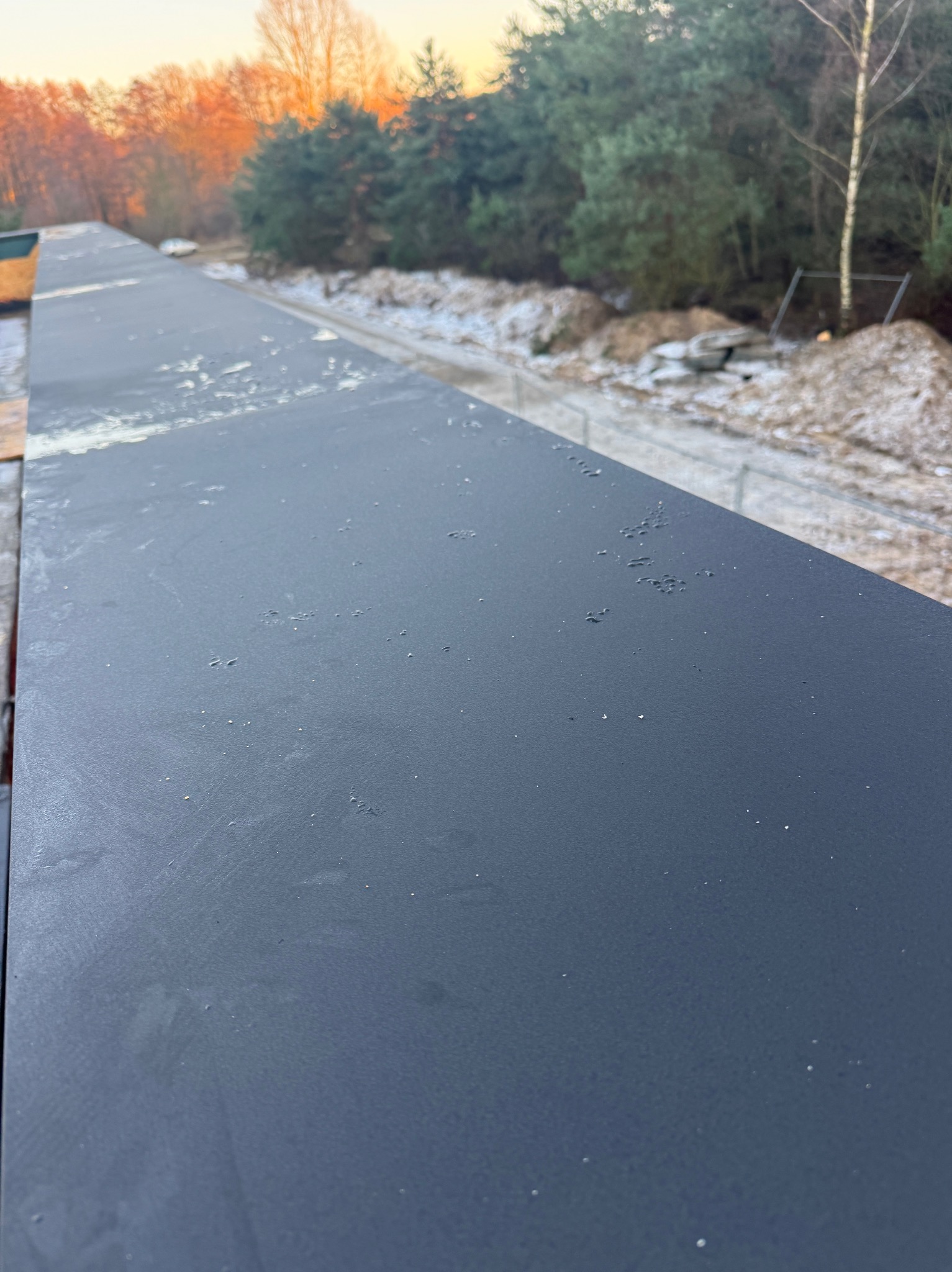 Szczegółowe ujęcie nowej, matowej obróbki blacharskiej na dachu, z widocznym tłem drzew i fragmentem nieba o zachodzie słońca. Minimalistyczny kadr.